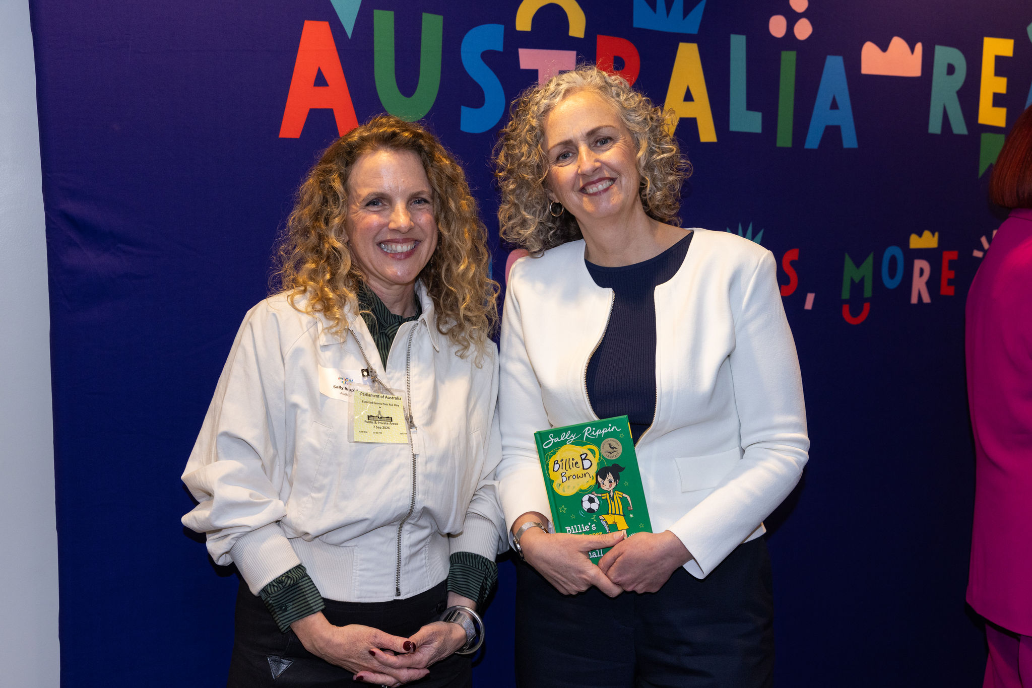 Two women smiling in front of a colourful Australia Reads banner at Parliament House, one holding a Billie B Brown children's book